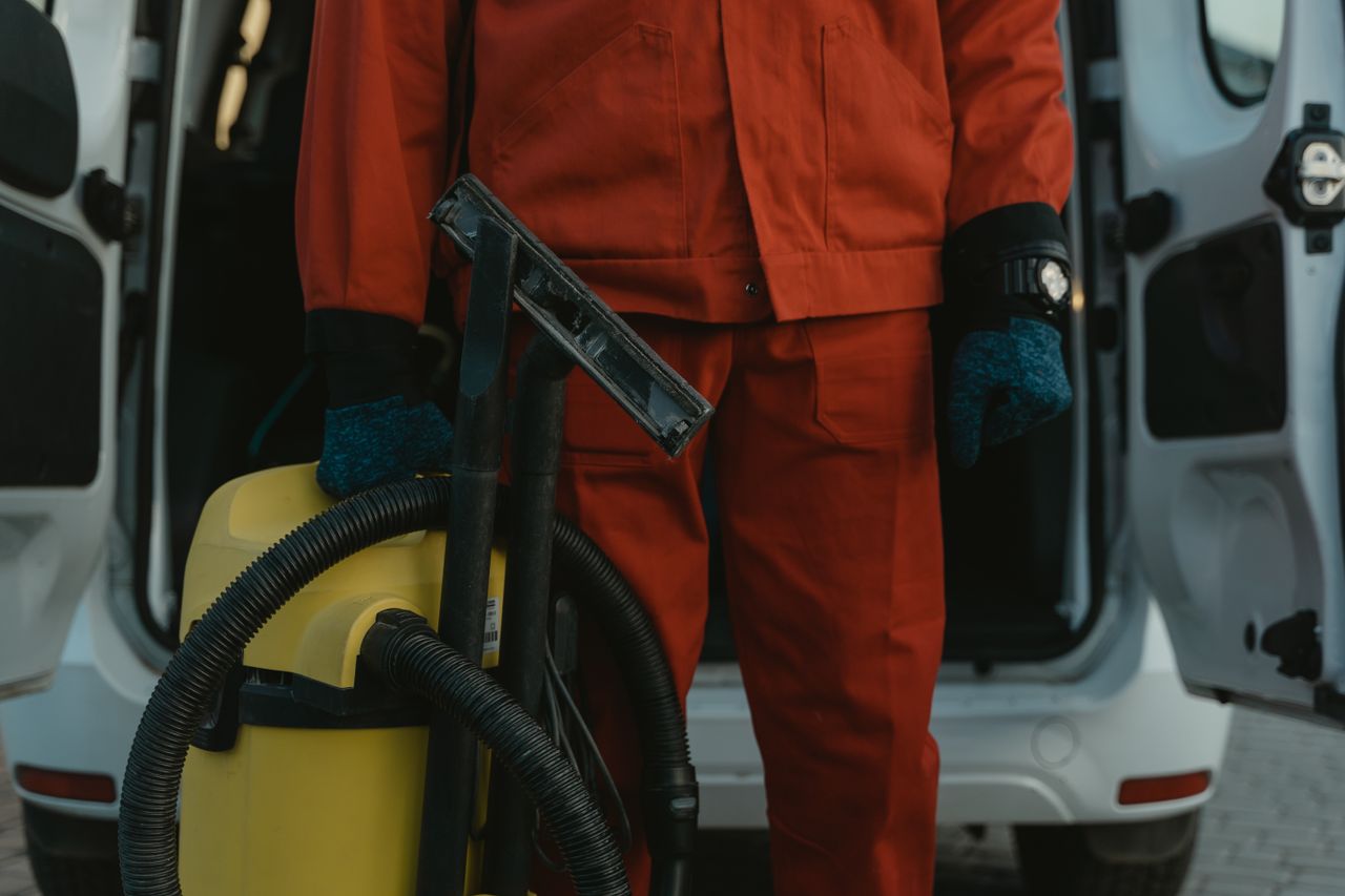 why-choose-us-02 A professional cleaner in a red uniform holding a vacuum beside a van, prepared for service.